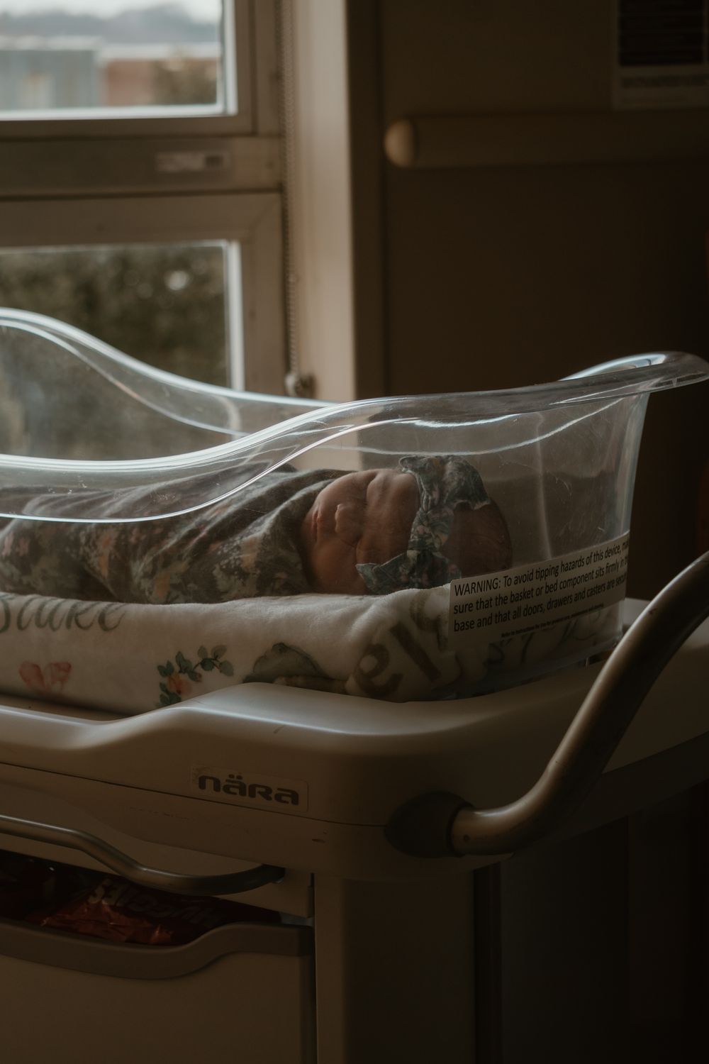 Newborn baby sleeping peacefully in a Nara hospital bassinet near a window in soft natural light.