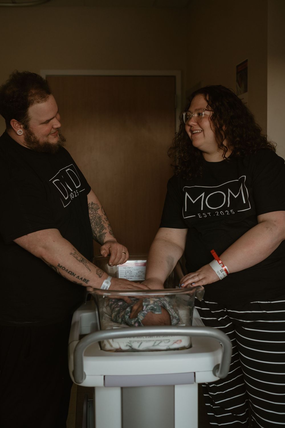 New parents wearing matching Mom and Dad black t-shirts smiling over their newborn in hospital bassinet.