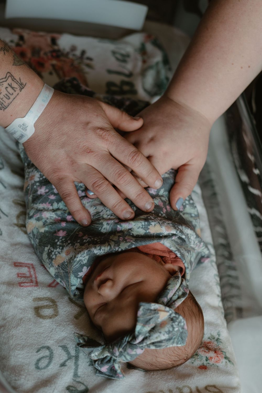 Tattooed parent gently placing hand on swaddled newborn baby wearing floral bow headband in bassinet.