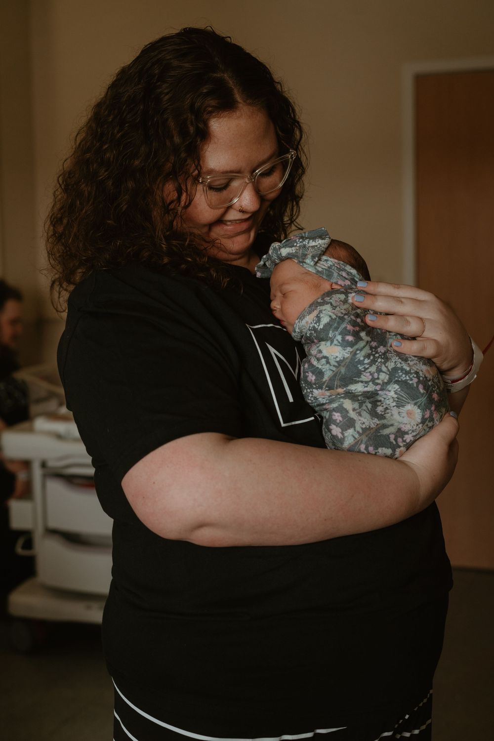 New mother in black Mom t-shirt lovingly cradling her swaddled newborn baby in hospital room.
