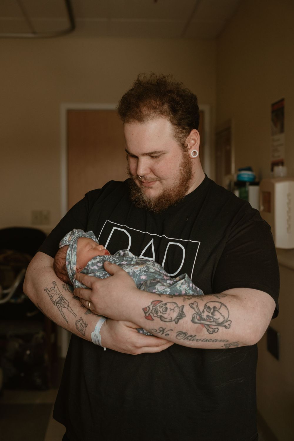 Tattooed new father in black Dad t-shirt tenderly holding his swaddled newborn baby in hospital.