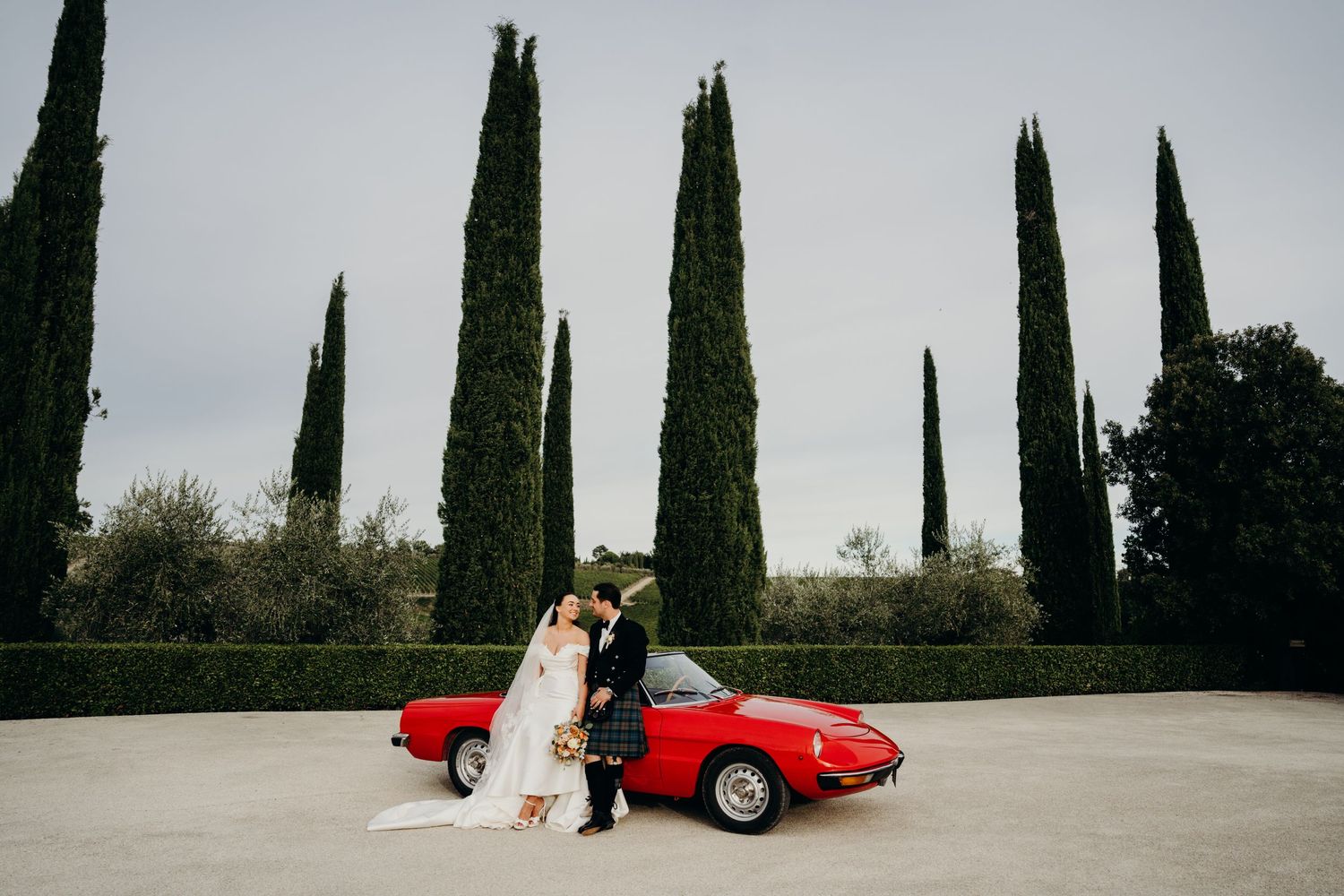 Wedding couple poses beside a classic red sports car with tall cypress trees in background.