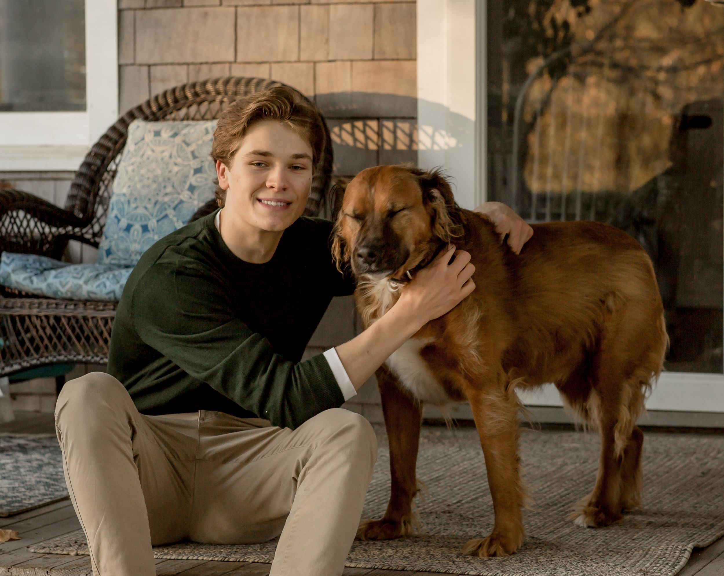 Young man crouching beside a large fluffy brown dog on a porch, smiling warmly.