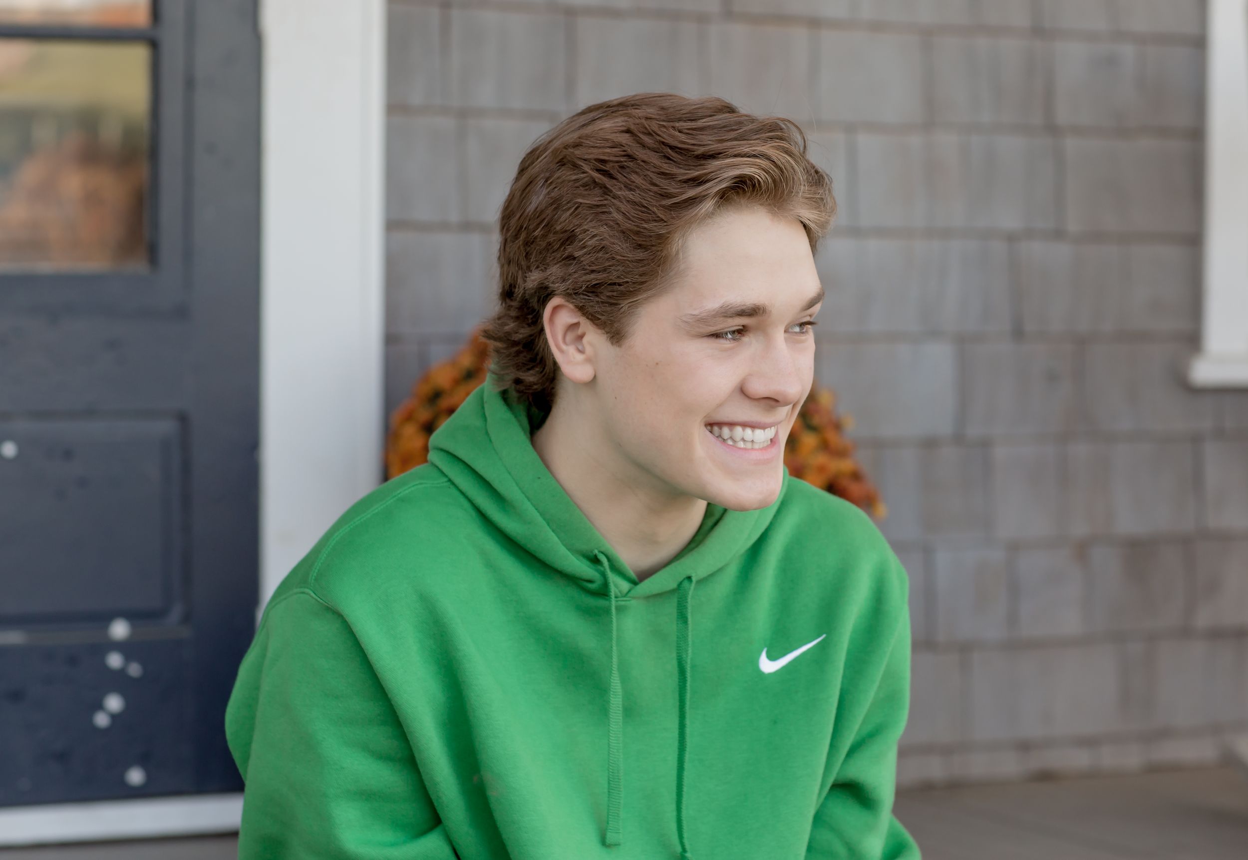 Smiling young man in a green Nike hoodie sitting on a porch, looking to the side.