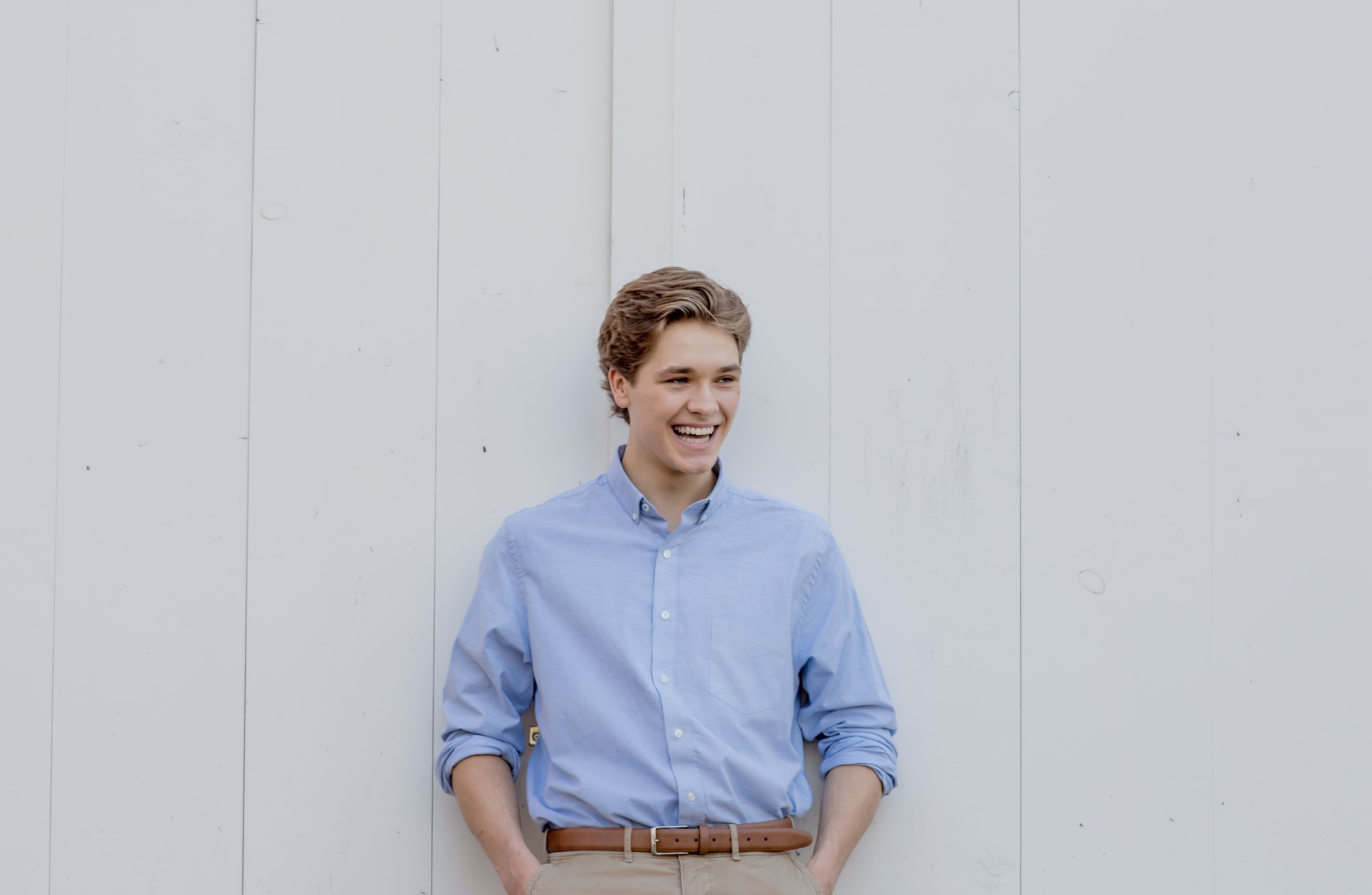 Smiling young man in a light blue button-up shirt standing against a white wall outdoors.