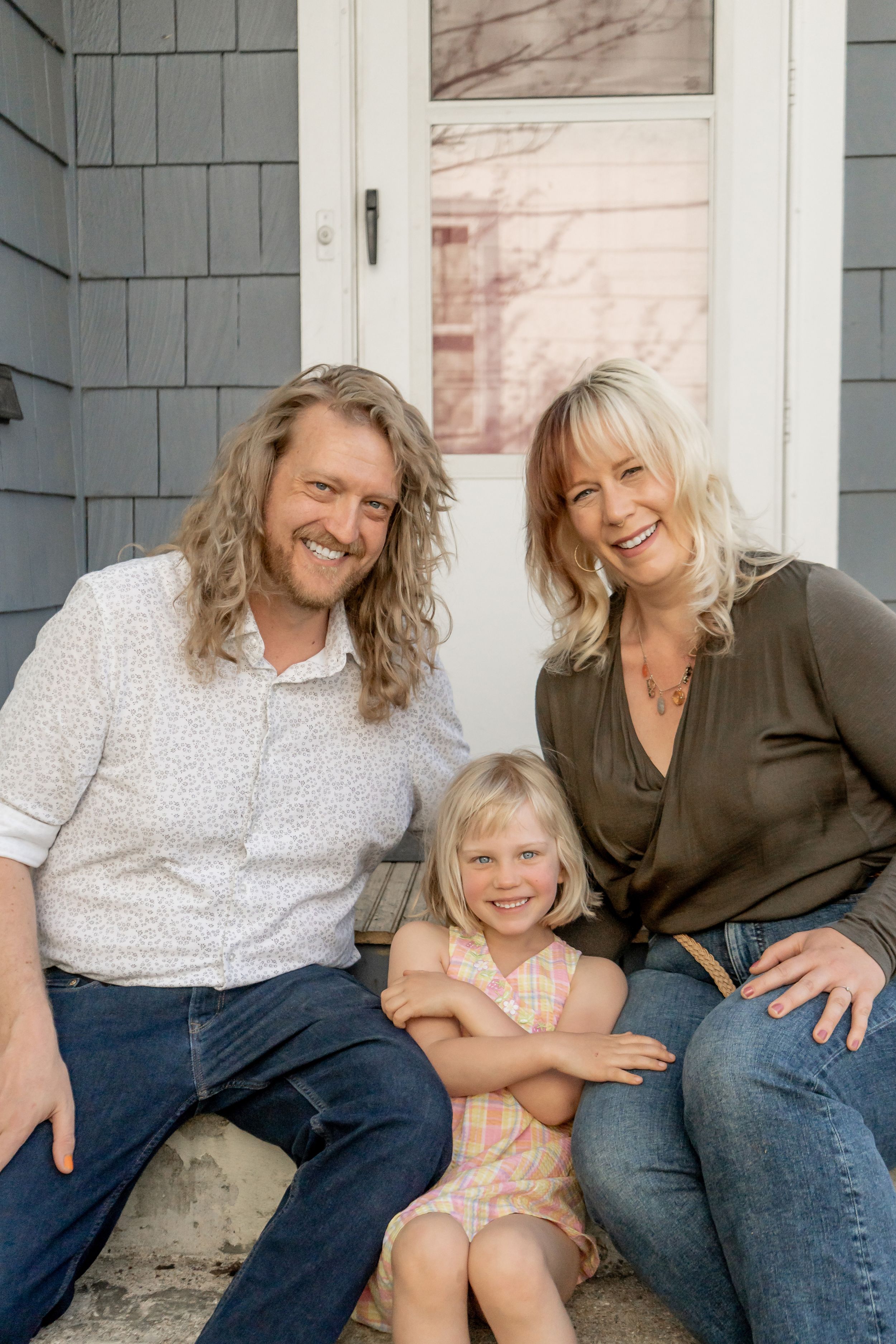 Smiling family of three, two adults and a young blonde girl, posing on front porch steps.
