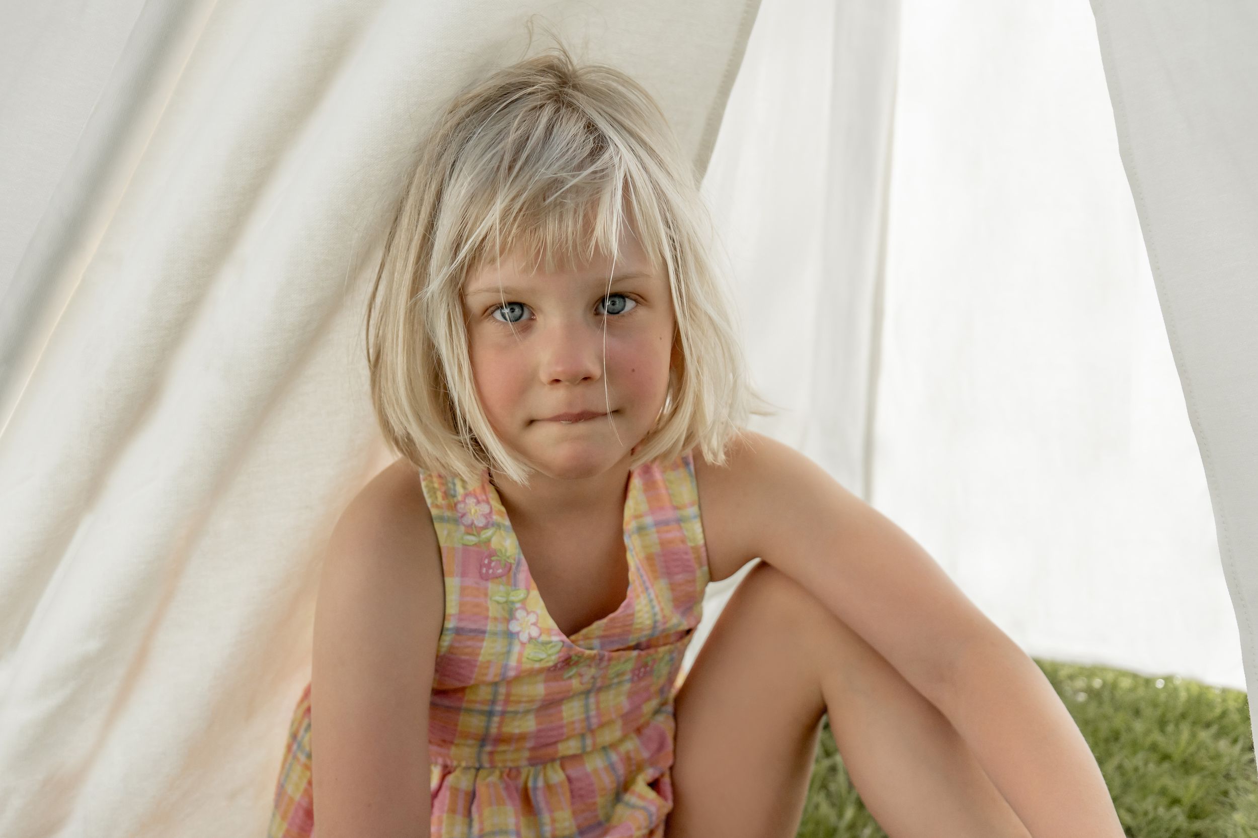Young blonde girl in a plaid dress sitting quietly inside a white teepee tent.