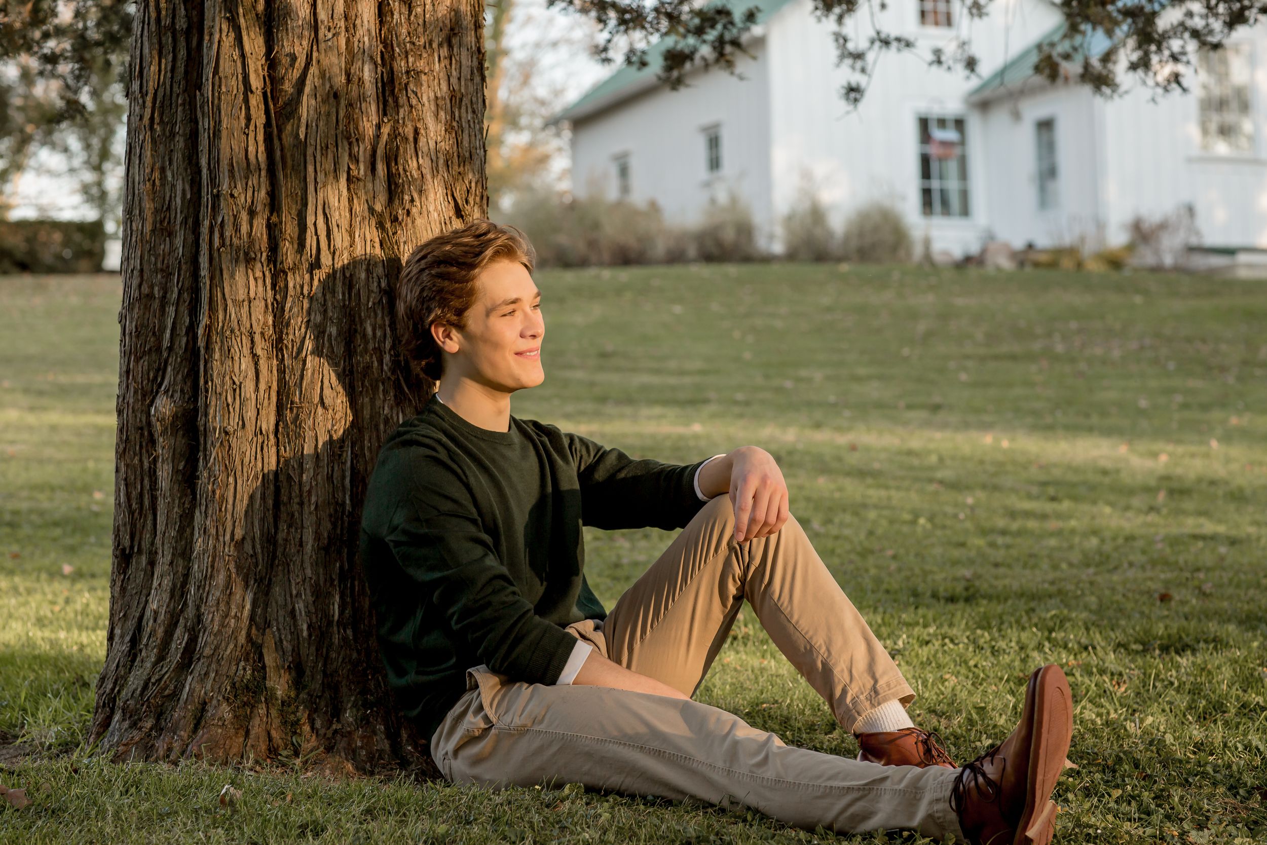 Young man sitting against a tree trunk in a green field, gazing into the distance.