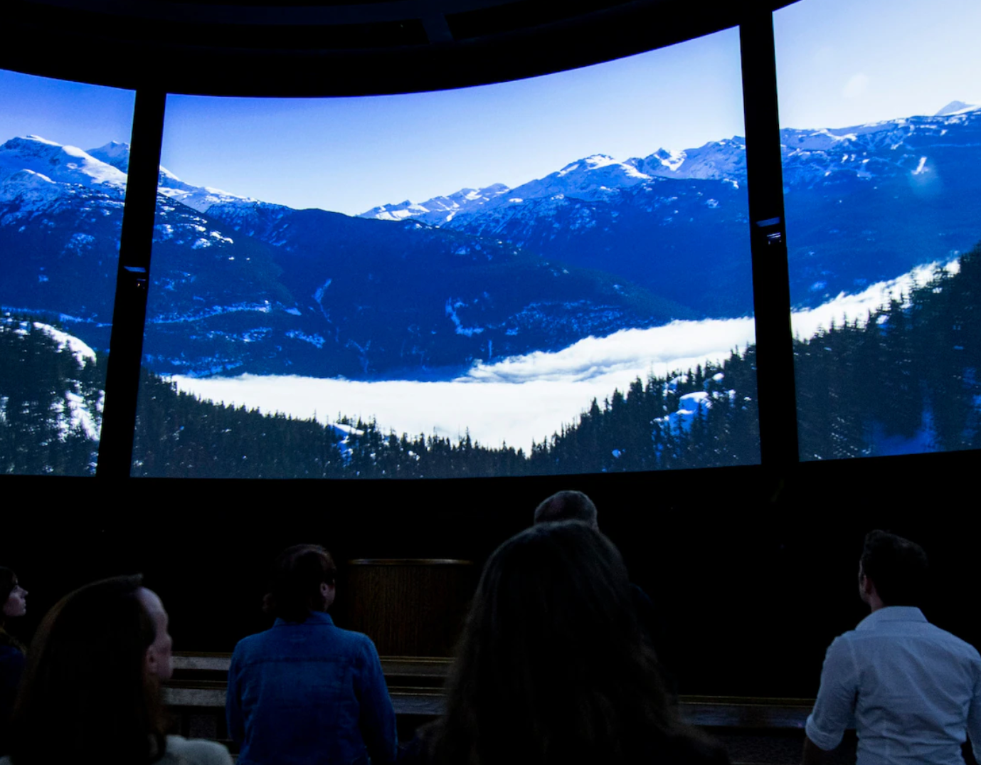 Theater audience viewing panoramic mountain landscape with snow covered peaks on curved screen.