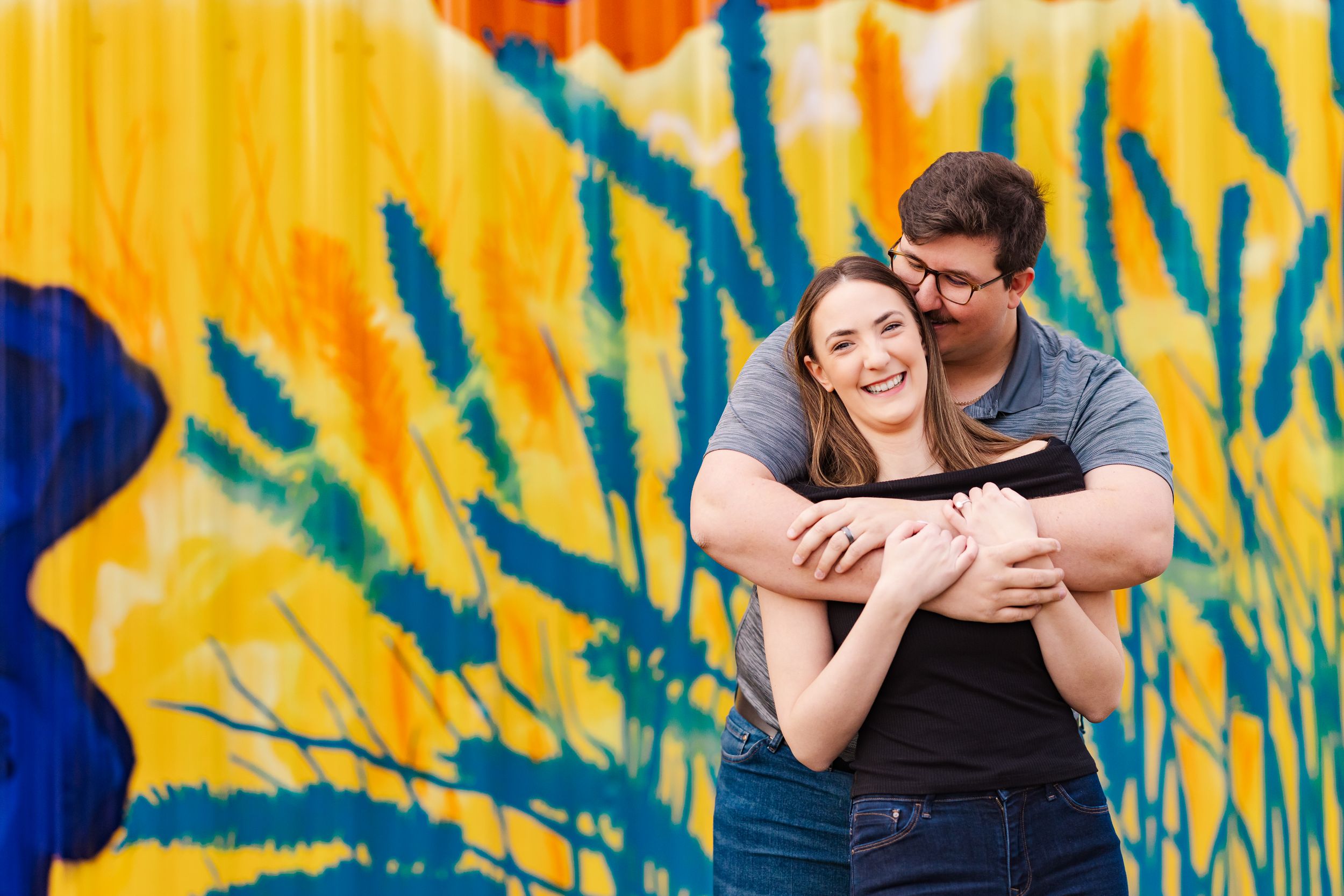A couple embraces and laughs together in front of a vibrant yellow and blue graffiti wall.