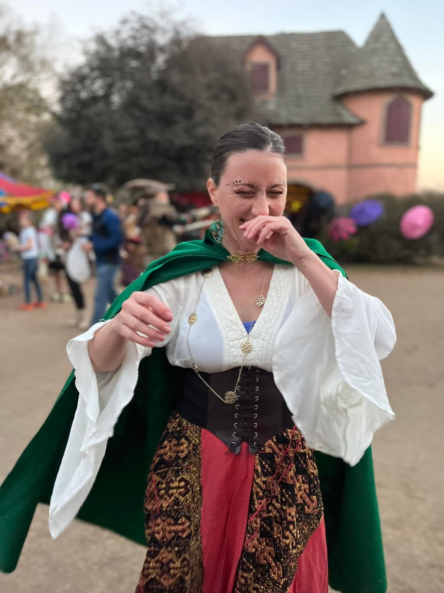 Woman in medieval renaissance faire costume wearing white blouse, corset and green cloak stands outdoors near buildings.