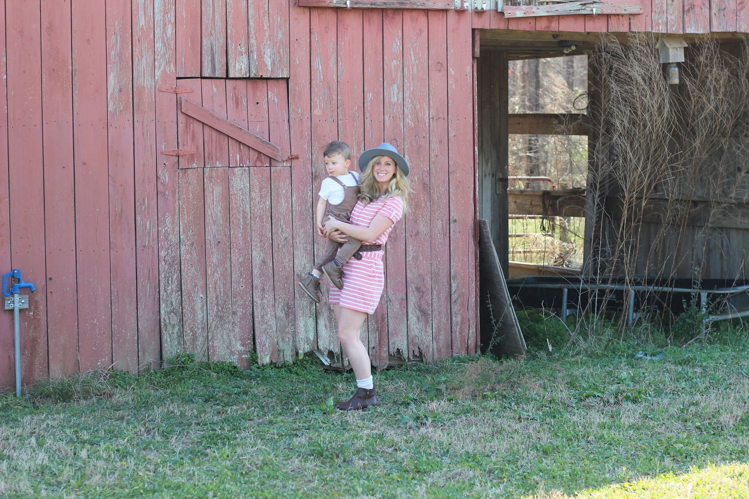 Someone wearing a pink dress stands in front of a rustic red barn wall on a grassy lawn.