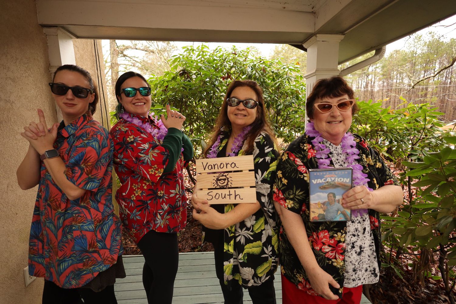 A group wearing colorful Hawaiian-style shirts poses together on a covered porch with tropical plants in the background.