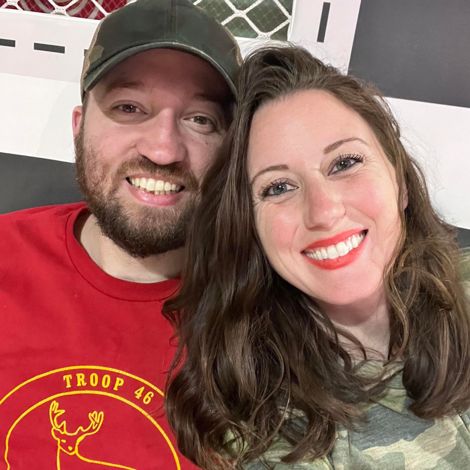 Happy couple taking selfies outdoors wearing casual attire including a baseball cap and camo jacket.