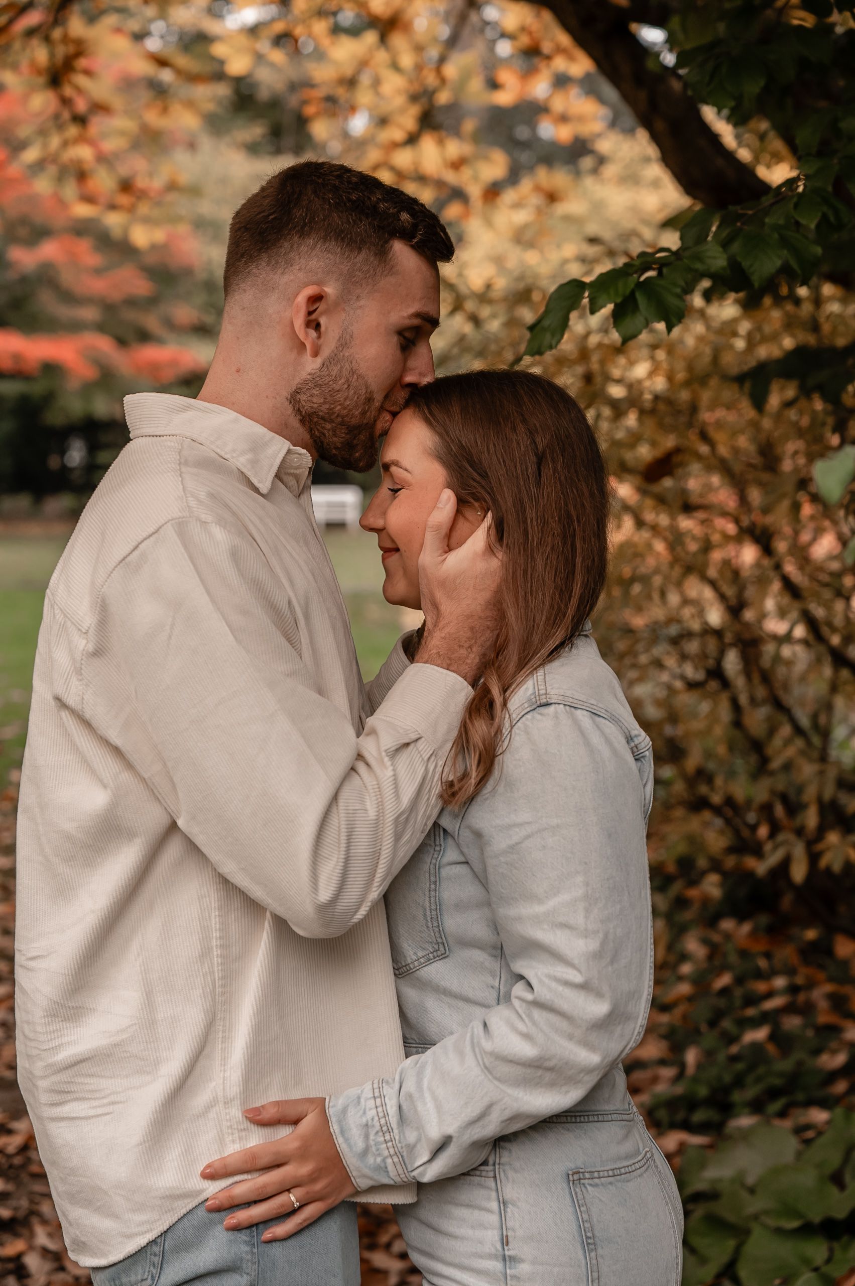 Couple in matching white outfits embrace tenderly among autumn trees during romantic outdoor photoshoot.