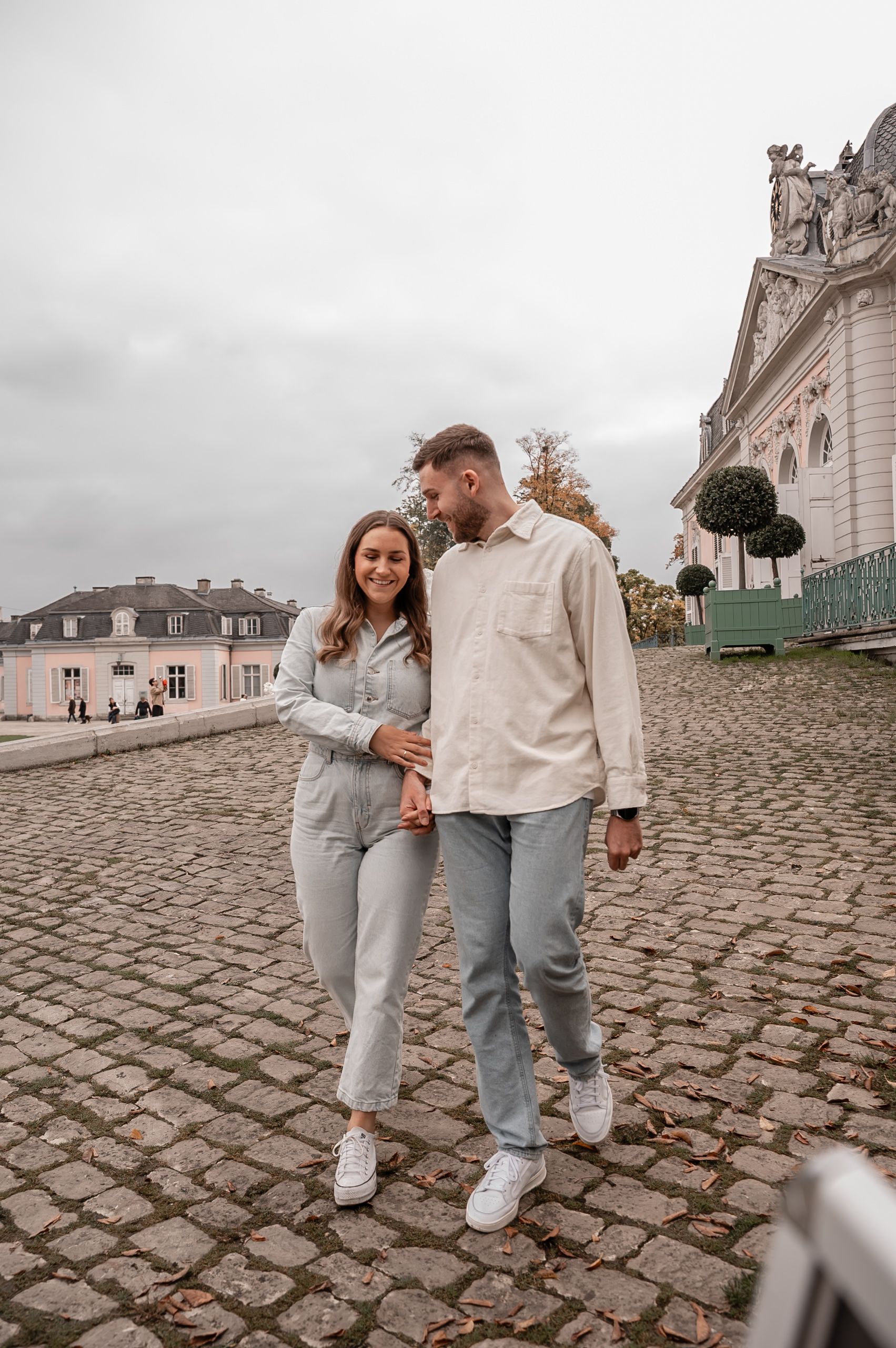 A couple in casual attire walking hand in hand on cobblestone pavement near historic buildings.