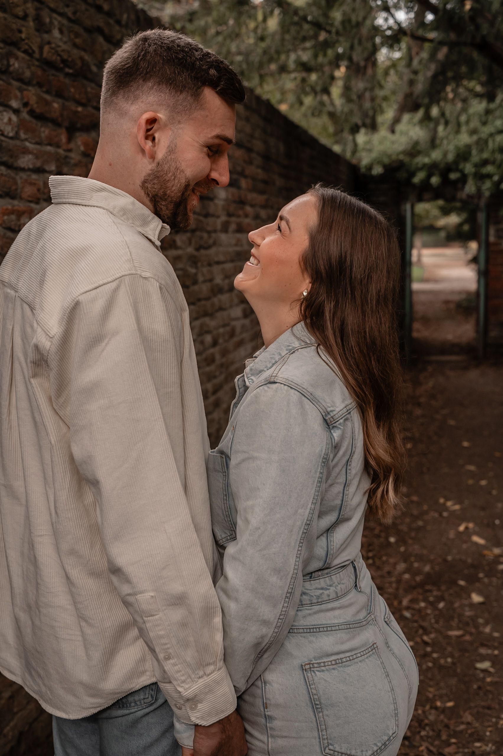 A romantic couple laughing together against a brick wall in a park setting with natural lighting.