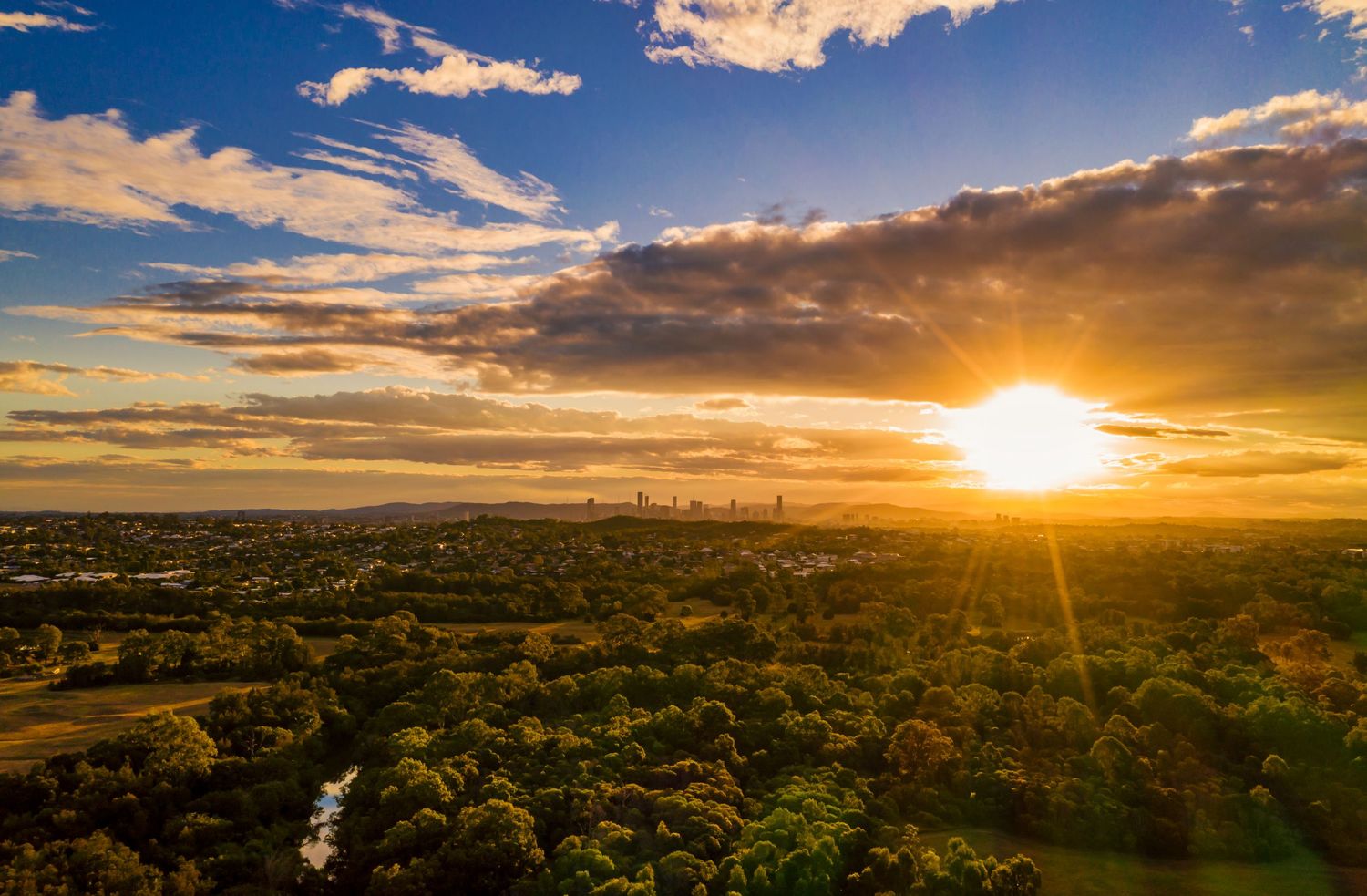 Vibrant sunset over urban landscape with river meandering through green spaces and distant city buildings.