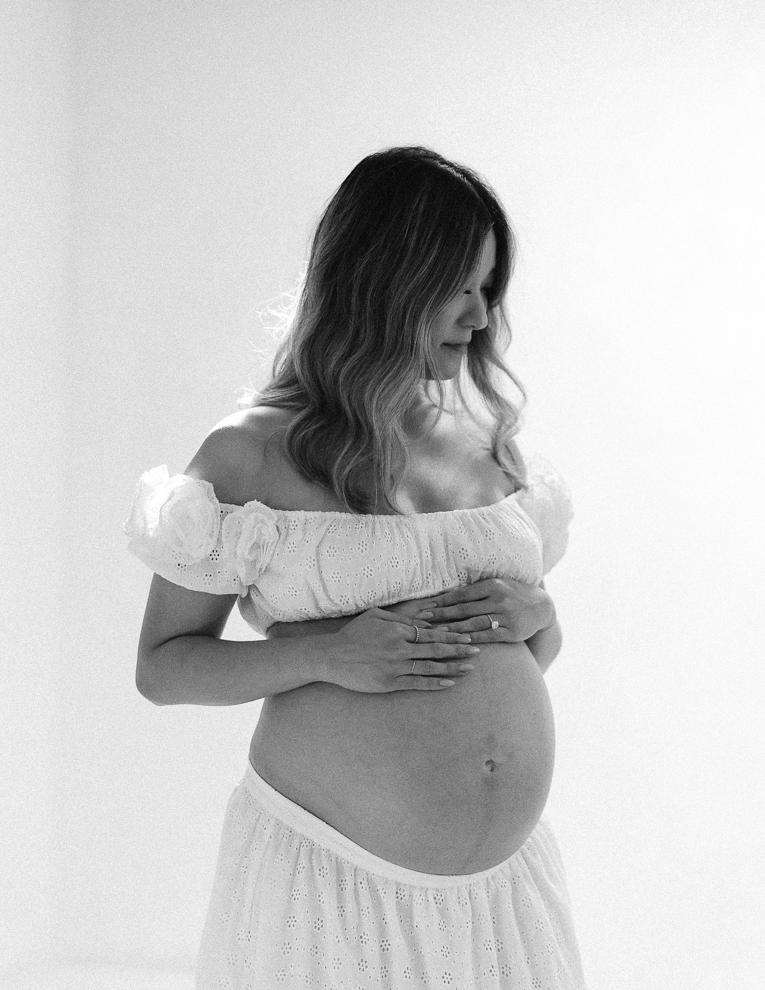 Black and white maternity photo showing expectant mother in off-shoulder white dress tenderly holding her baby bump.