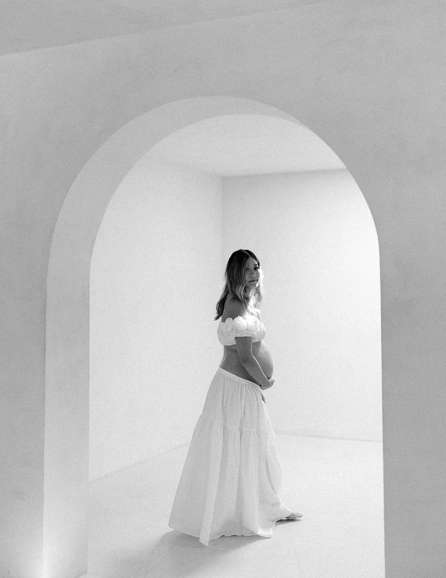 A figure in a white dress moves through a minimalist white curved archway corridor creating an ethereal atmosphere.