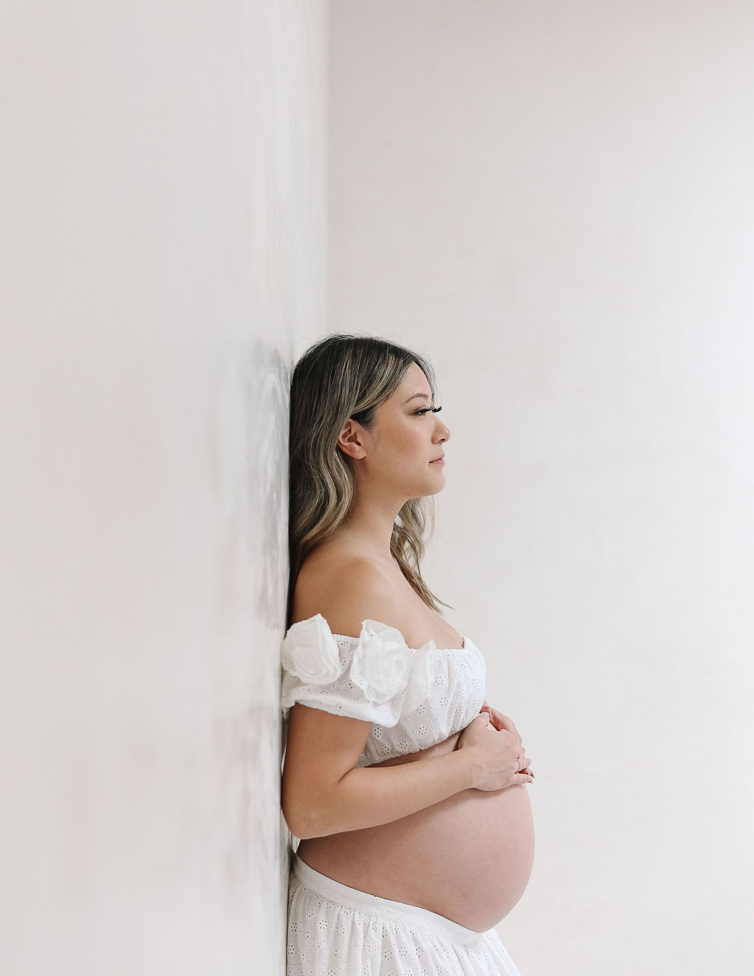 Maternity portrait in white dress against white wall showing profile view and bare shoulders in minimalist style.