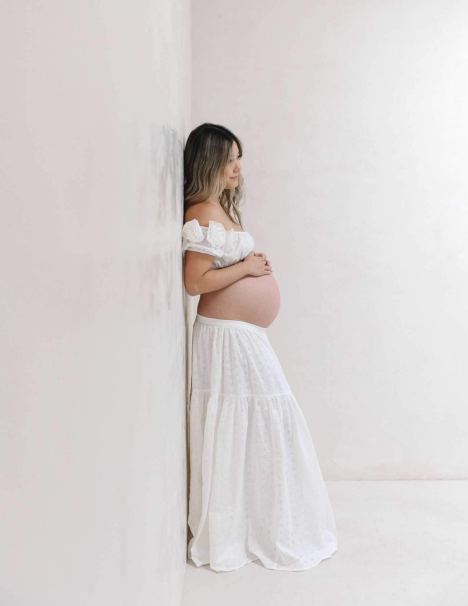 A pregnant woman in a flowing white off-shoulder dress stands against a plain wall in a minimalist maternity photo.