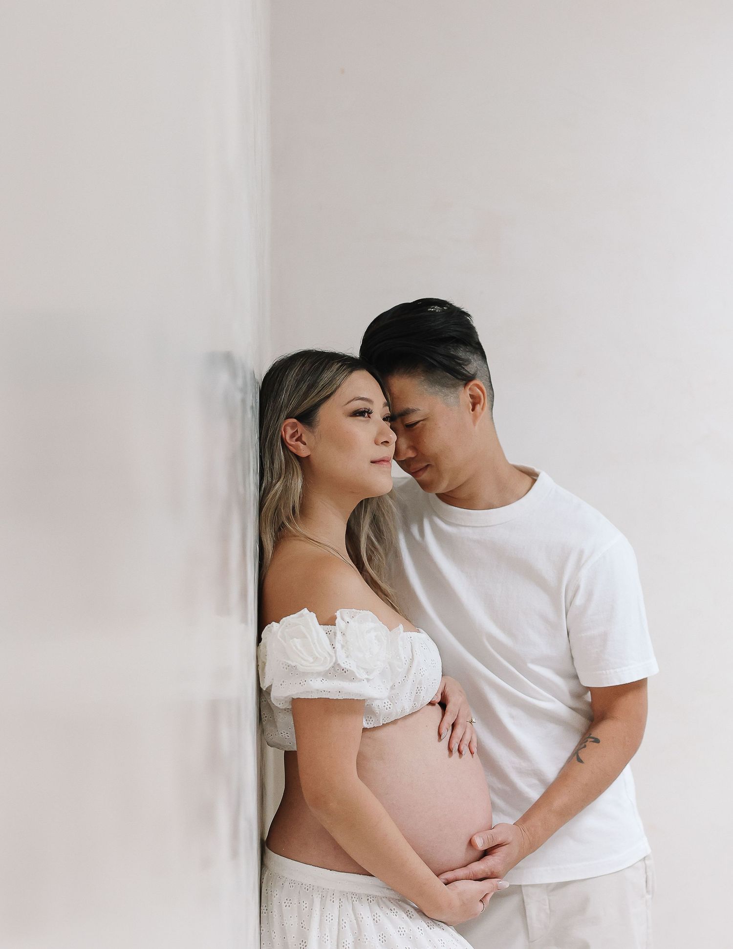 A couple in white outfits shares an intimate maternity photo session against a minimalist light background.