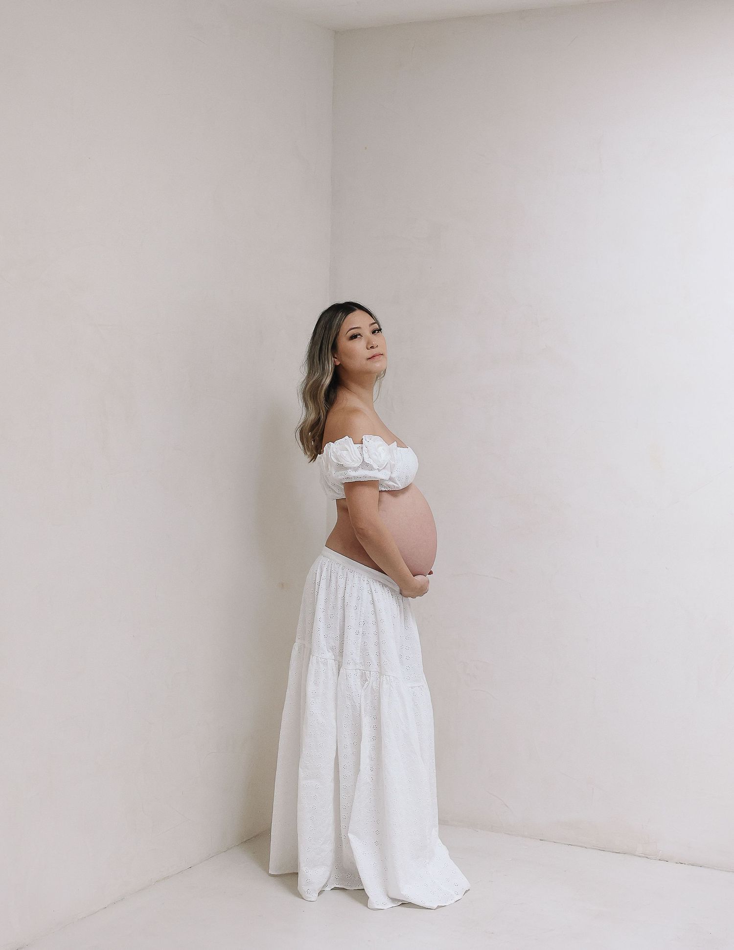 Ethereal maternity portrait in white off-shoulder dress against minimal white corner background.