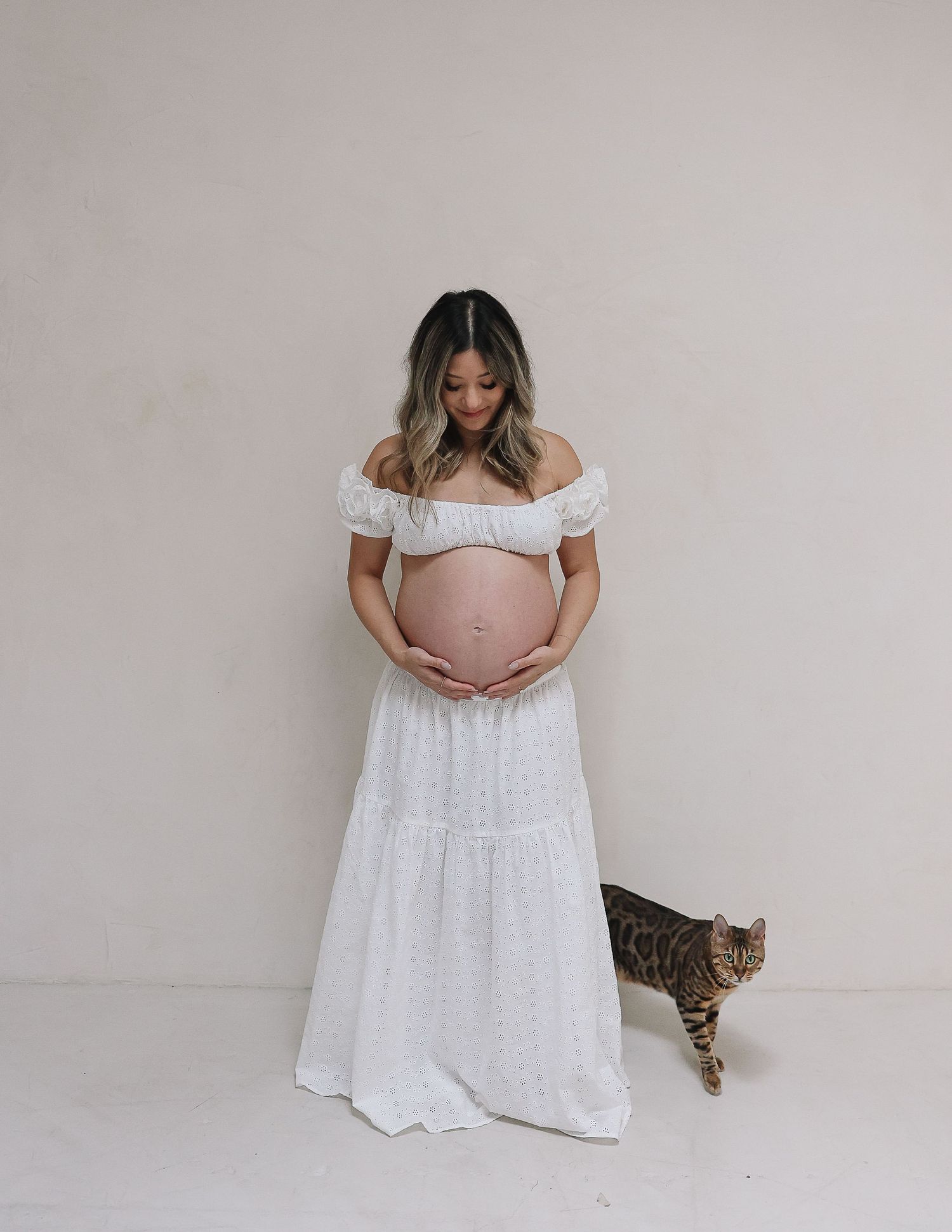 Expectant woman in white off-shoulder dress poses against neutral wall while tabby cat walks by in studio maternity photo.