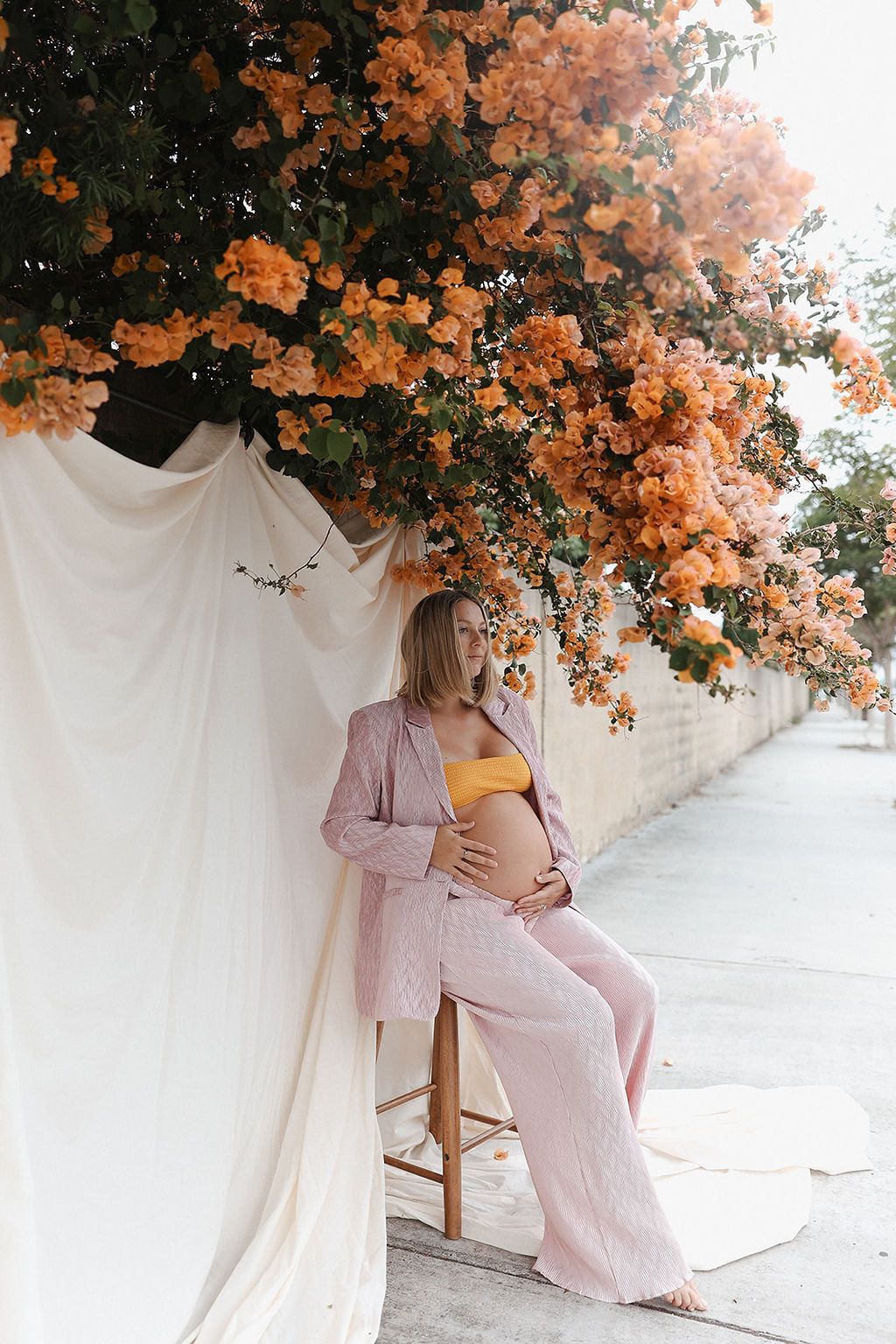Elegant maternity photo under blooming orange bougainvillea with soft pink accents and draped white fabric.