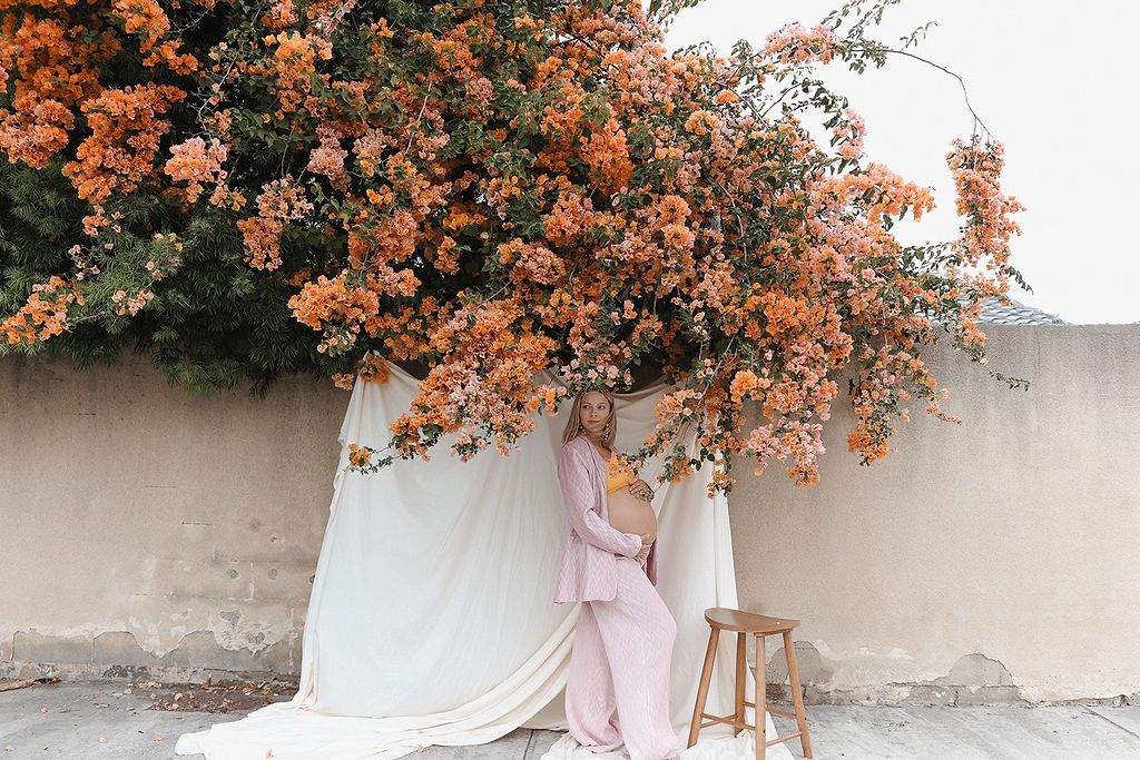 A dreamy scene with a wooden stool beneath a blooming coral-colored bougainvillea against a white backdrop and stone wall.