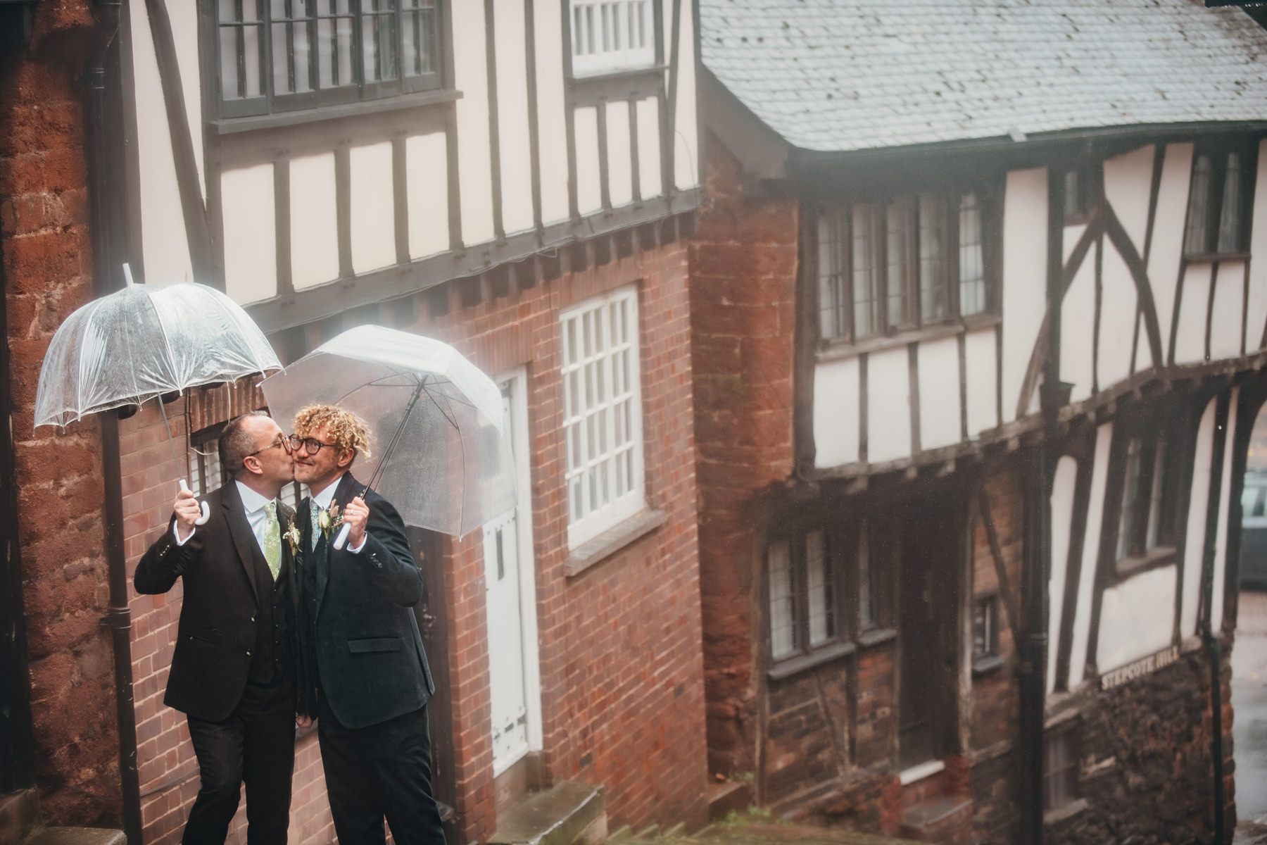 Two people in formal attire share umbrellas while standing together on a rainy day near a Tudor-style brick building.