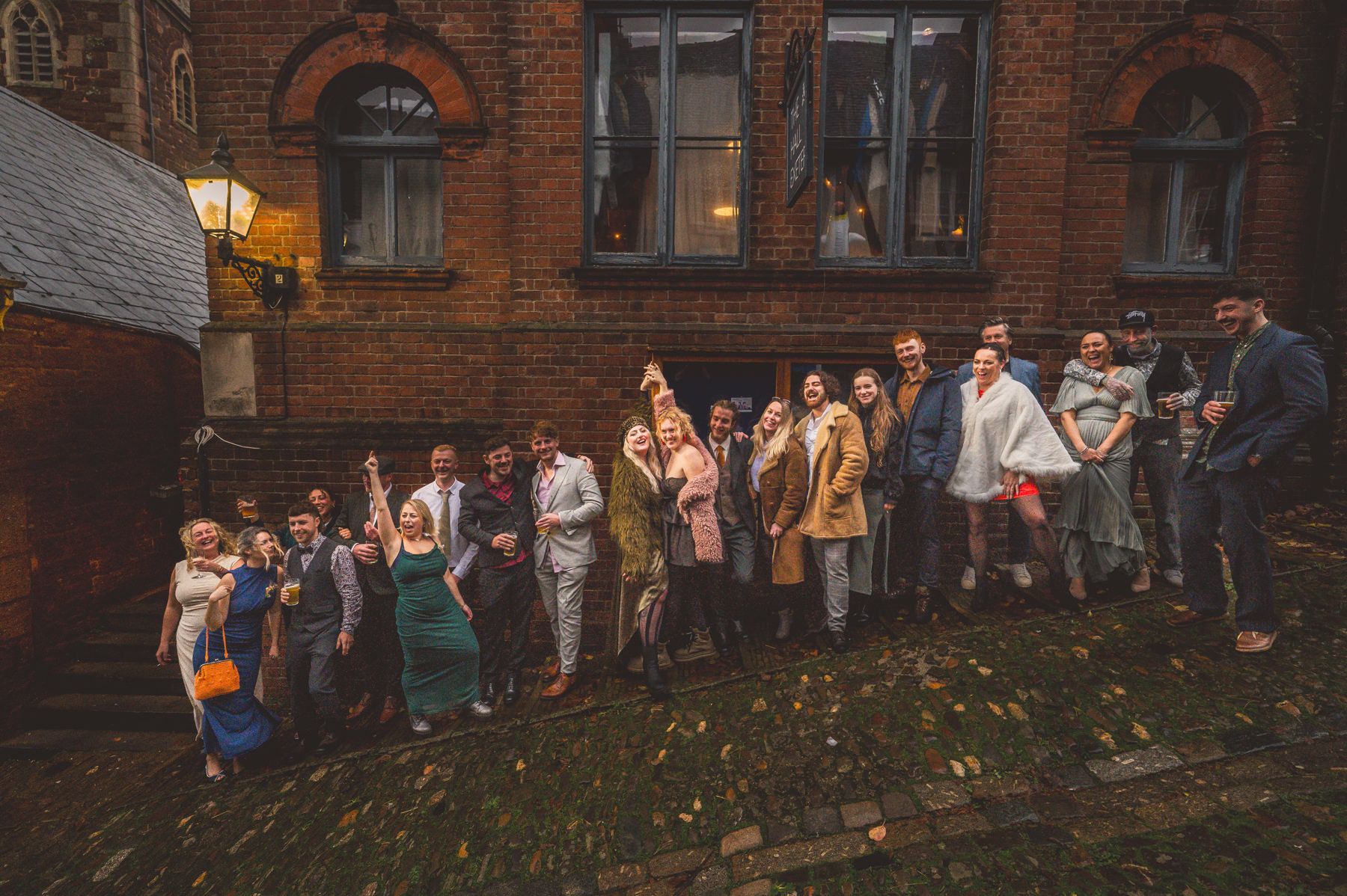 A large group of people gather outside a historic brick building at dusk with warm lighting in an autumn setting.