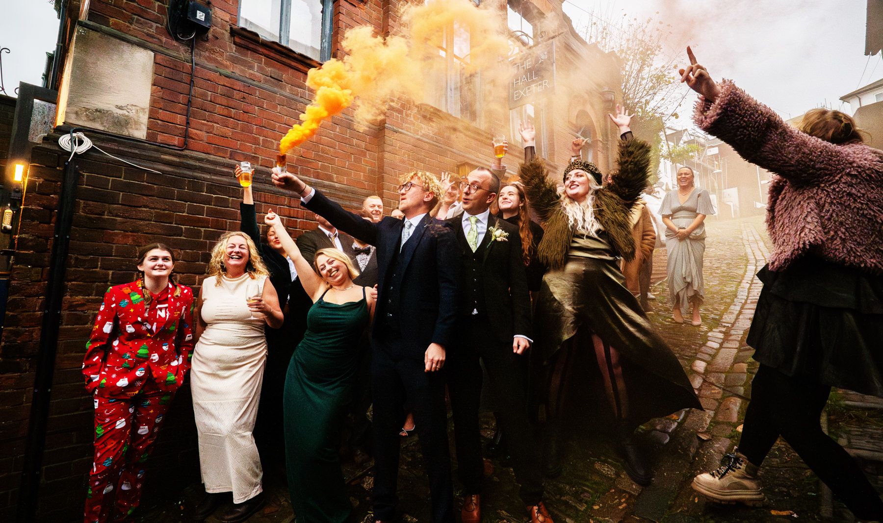 A wedding party celebrates with colorful smoke bombs outside a brick building, creating a festive and joyful atmosphere.