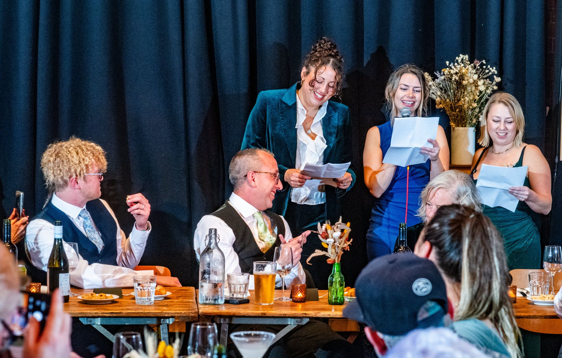 People gathered around a dining table during a celebratory event with drinks and laughter at what appears to be a reception.