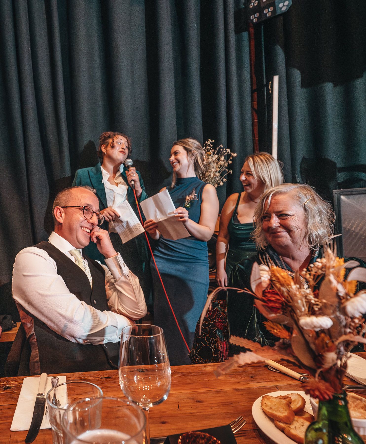 A group of people sharing drinks, laughter and conversation around a wooden table at a social gathering.