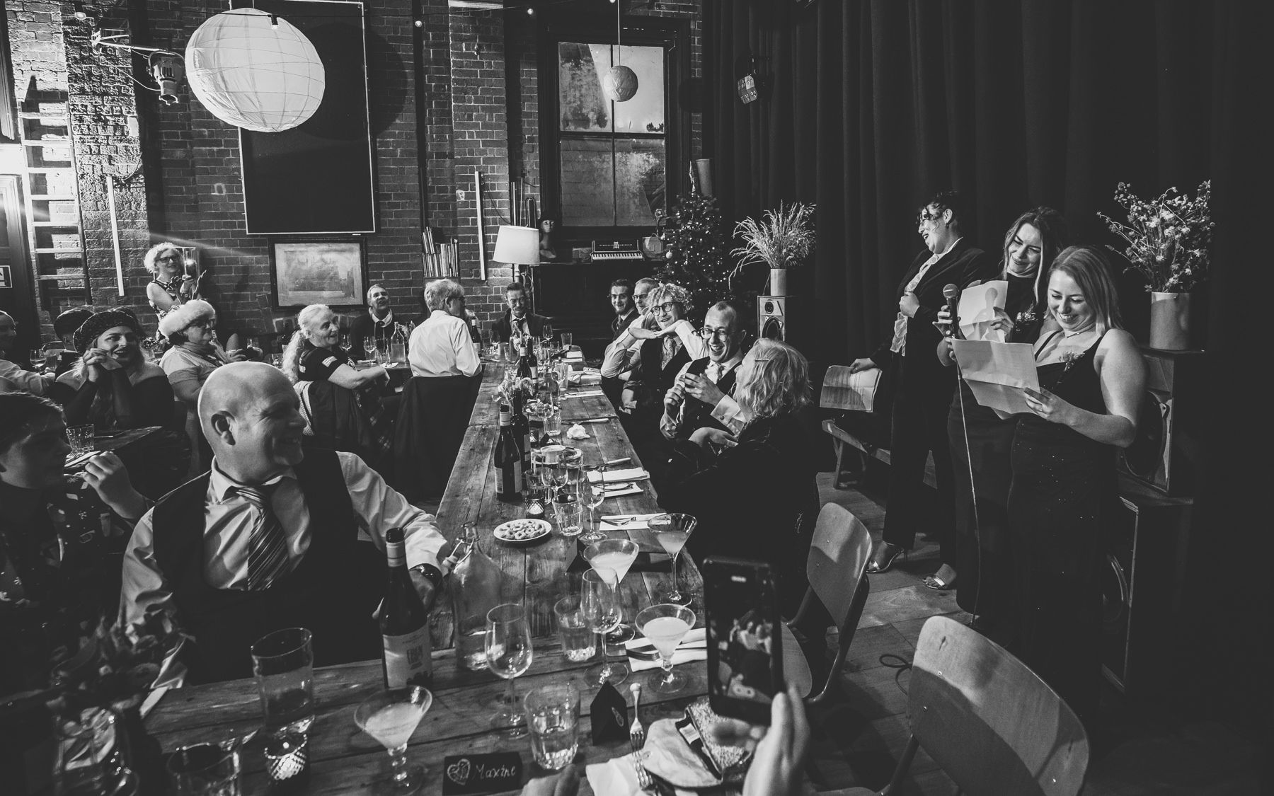 Black and white photograph of people dining and socializing at a long table in a dimly lit restaurant.