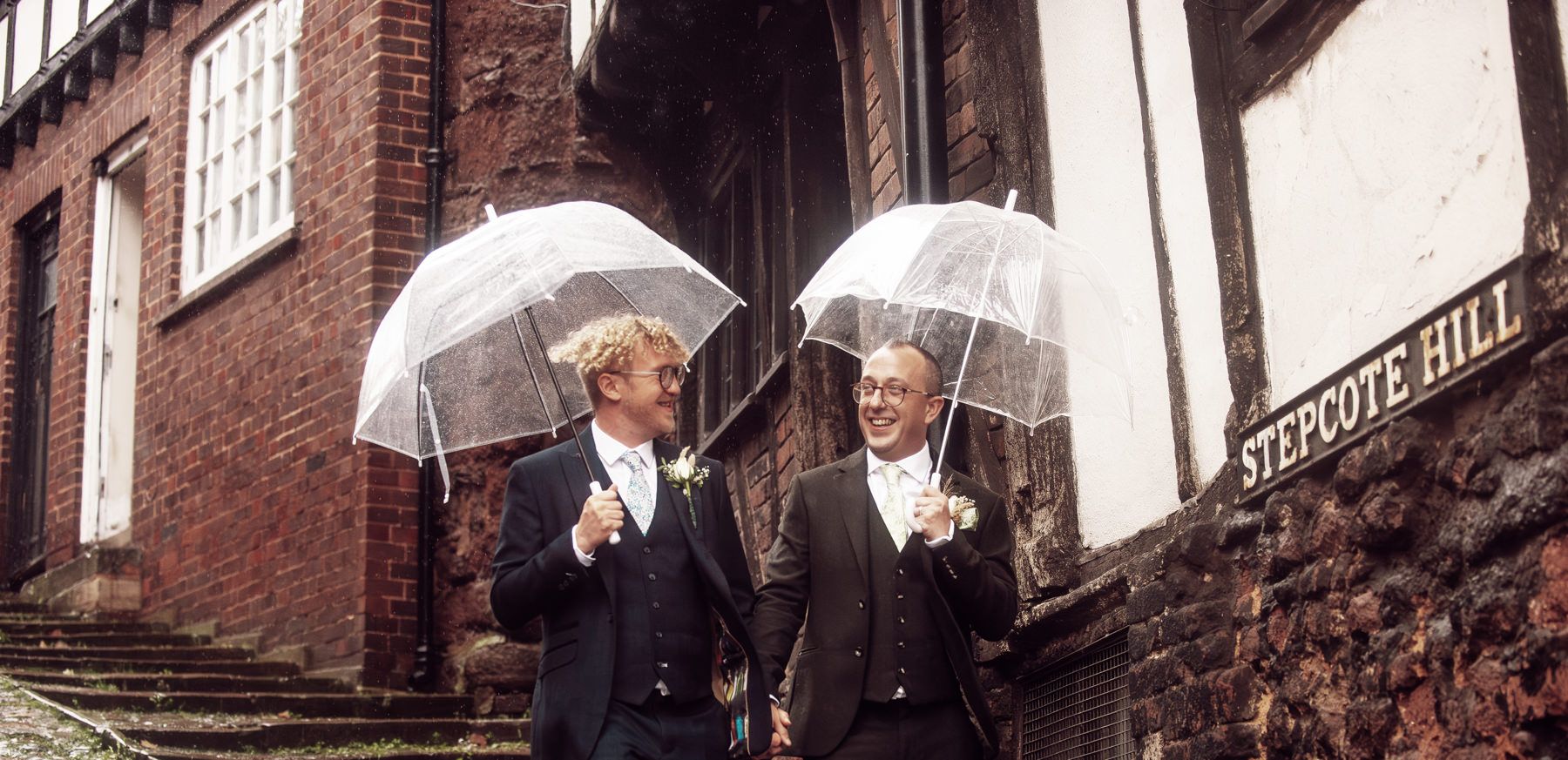 Historic cobblestone street with umbrellas and people walking past red brick buildings in the rain.