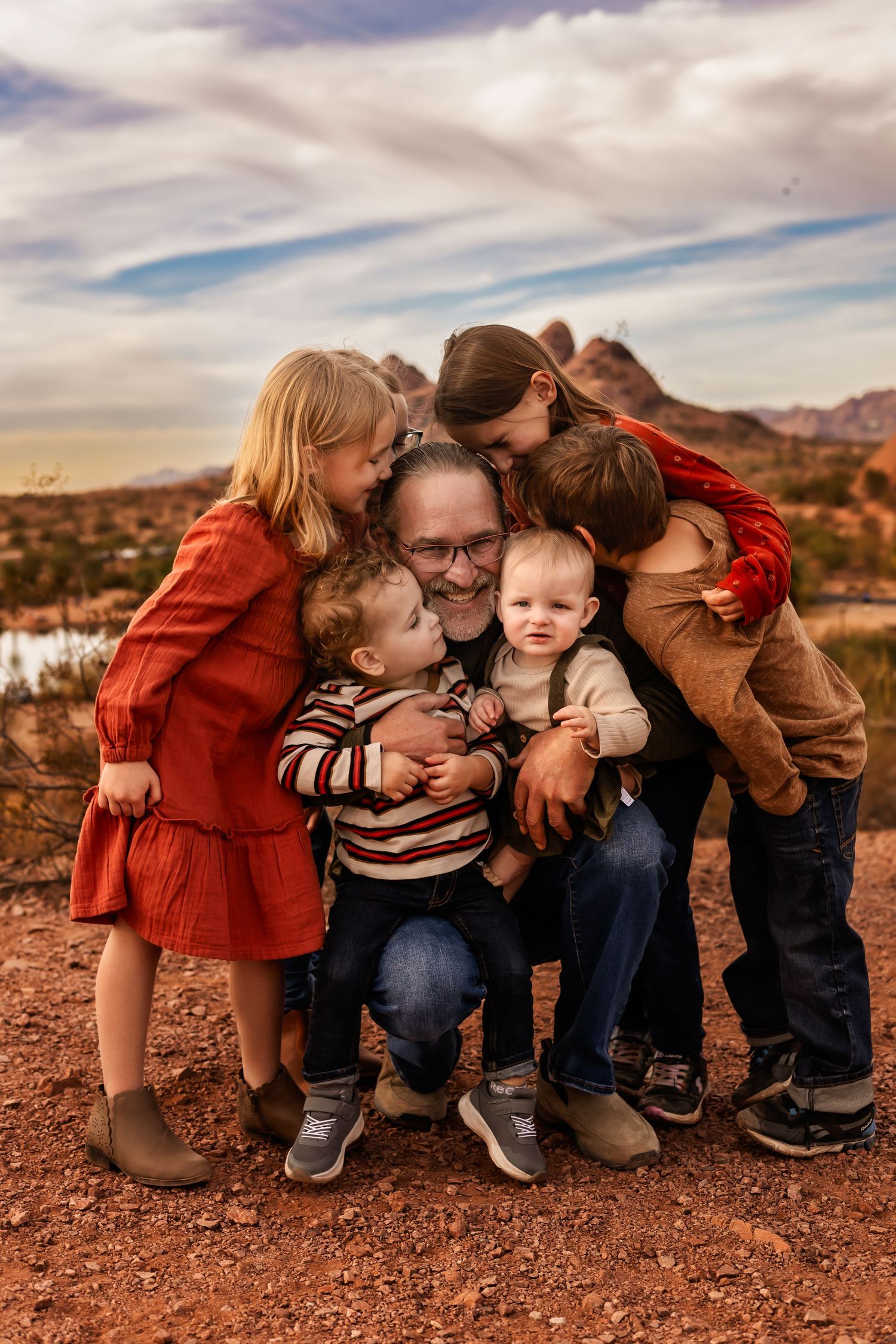 Family embraces together against desert mountain landscape in rustic red clay setting during sunset.