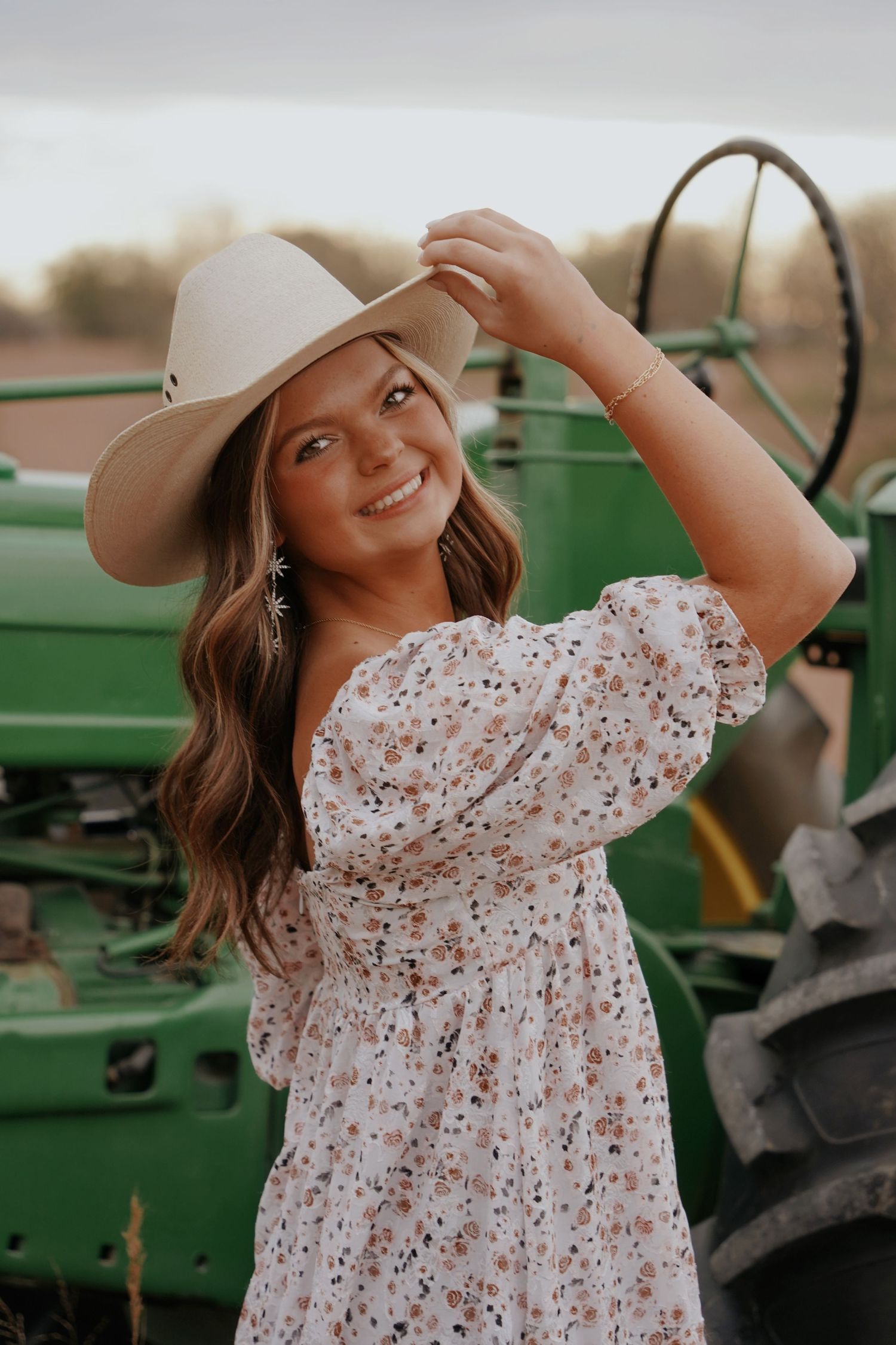 Smiling young woman in floral dress and cowboy hat posing near a green John Deere tractor at sunset.