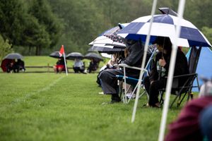 Golf spectators shelter under umbrellas during a rainy day at the course, with golf bags visible on the green grass.