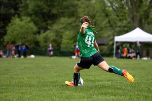 Youth soccer player in green jersey number 11 stretches to control the ball on a sunny grass field during a match.