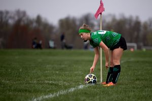 Soccer player in green uniform sets up a corner kick on grass field with a pink flag waving in the background.