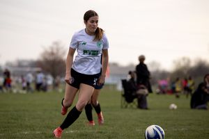 Soccer player in white jersey and black shorts dribbles ball during outdoor match on grass field with spectators.
