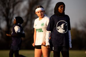 Soccer players stand on field during afternoon practice wearing white and black hoodies in warm sunlight.