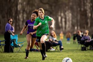 Youth soccer match action shows two players competing for the ball on a grassy field with spectators watching.