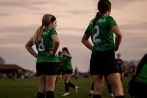 Female rugby players in green jerseys compete in a match during sunset on an outdoor field.