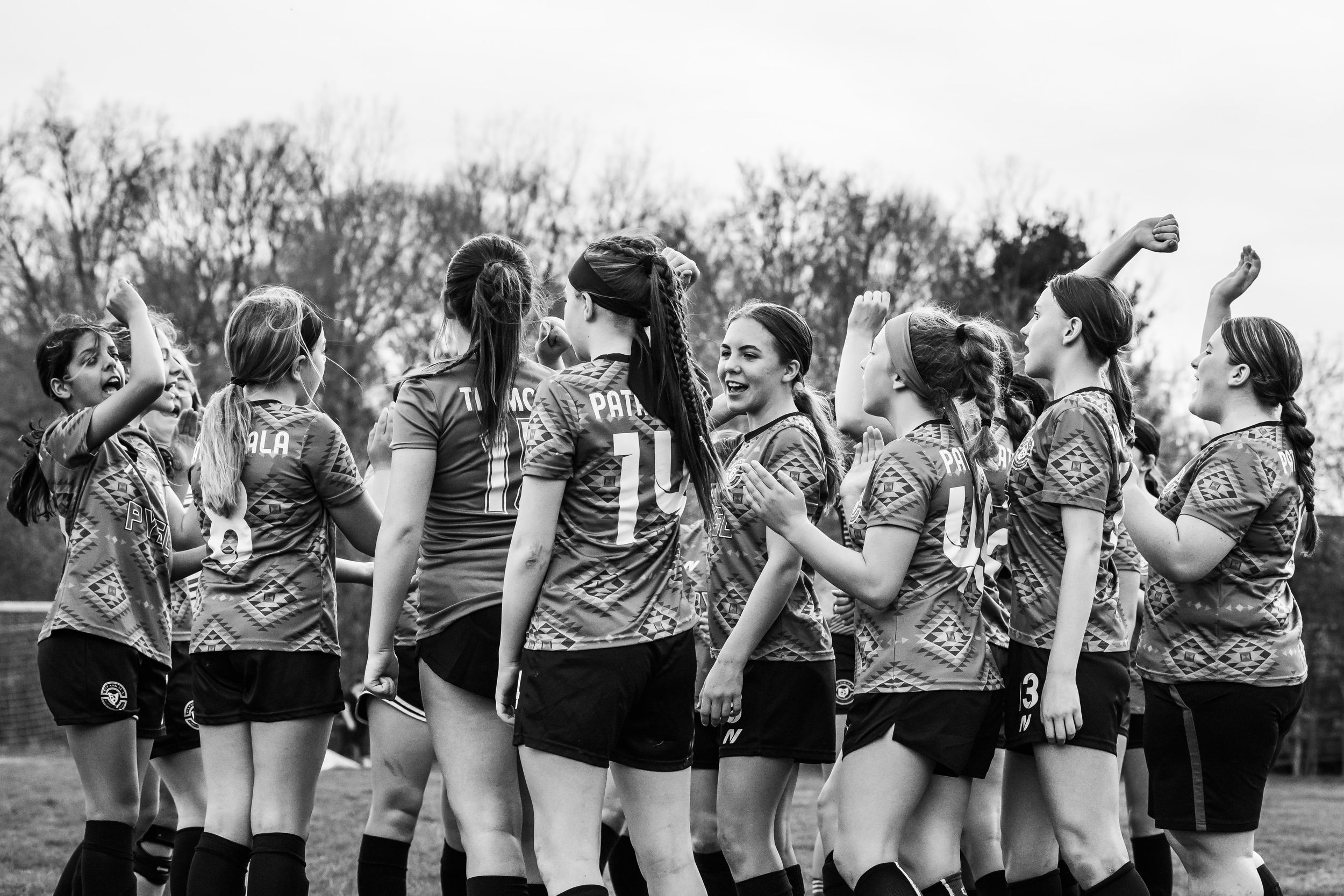 A girls sports team in matching jerseys huddles together on a field in black and white photography.