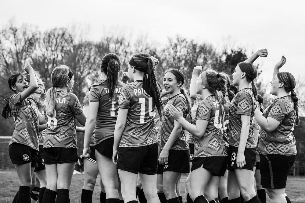 A girls sports team in matching jerseys huddles together on a field in black and white photography.