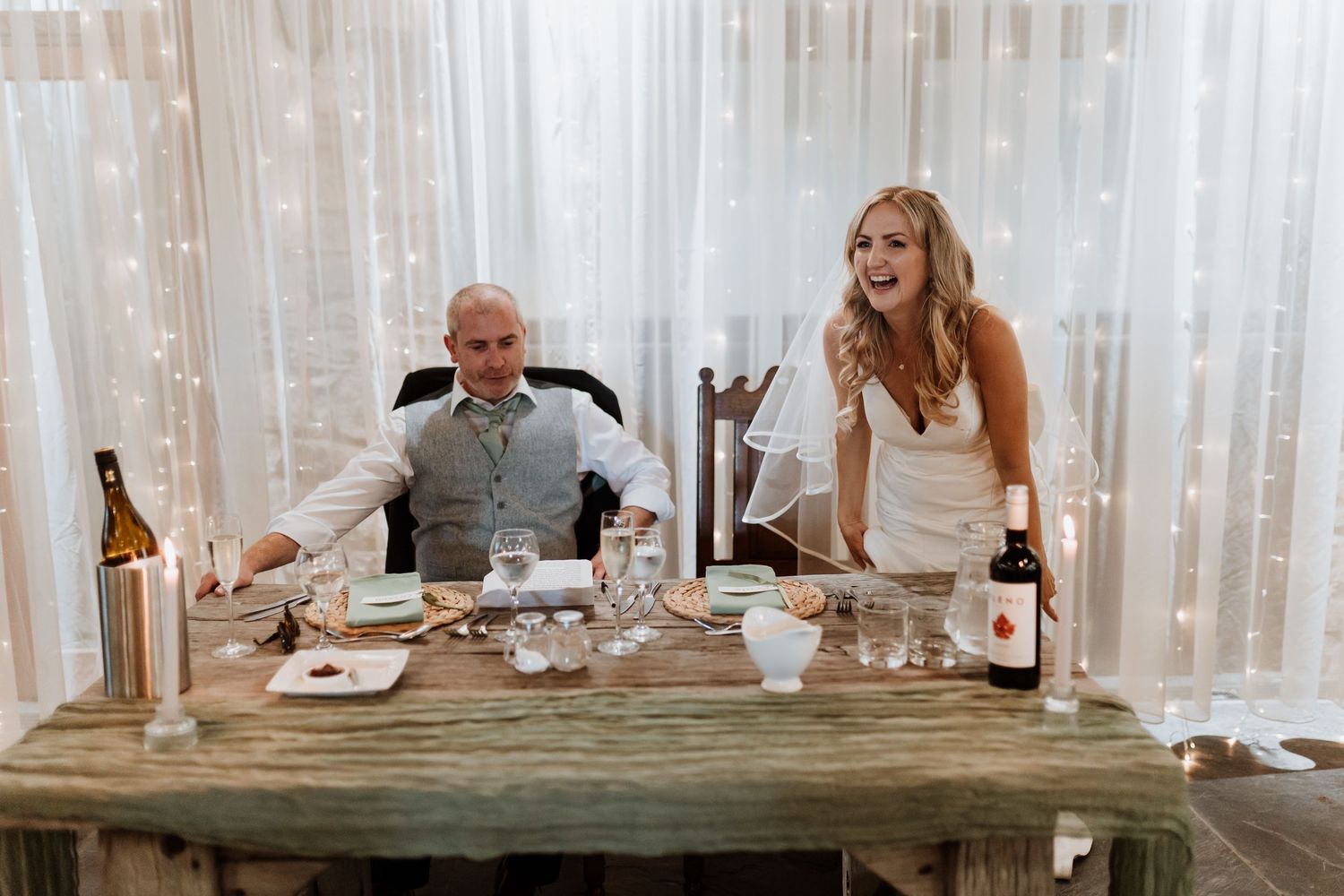 Wedding couple sharing a candid laugh during their dinner reception at a rustic wooden table with wine bottles.